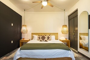 Une chambre à coucher avec un lit, deux lampes de chevet, un ventilateur de plafond, une table de chevet en bois, une porte sombre et un miroir sur le mur.