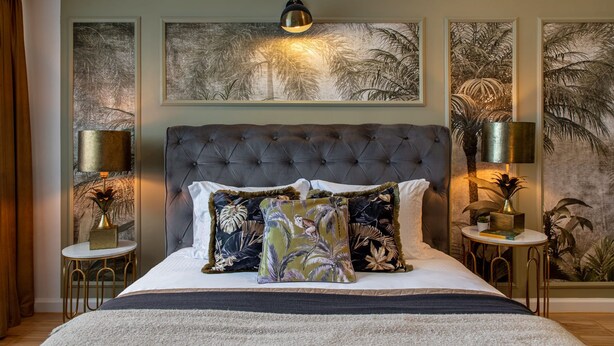 Une chambre à coucher avec une tête de lit capitonnée, un grand lit avec de nombreux oreillers, deux tables de chevet avec des lampes et des œuvres d’art encadrées accrochées aux murs.