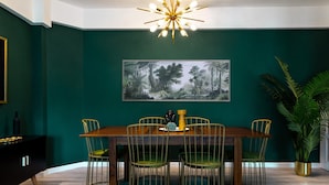 Uma sala de jantar com parede verde escura, uma grande pintura com tema de floresta, uma mesa de jantar de madeira e quatro cadeiras com almofadas verdes.