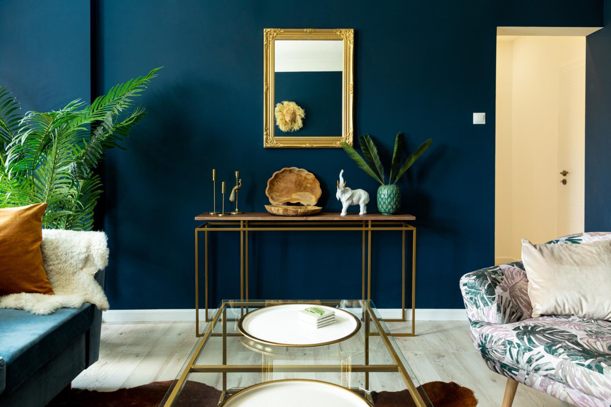 A room with a dark blue wall, a gold-framed mirror, a wooden console table with decorative items, a glass coffee table, a blue sofa, and a patterned armchair.