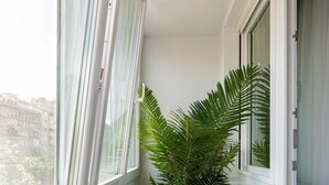 Uma varanda estreita com uma planta em vaso, paredes brancas e vista para edifícios.