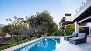 Piscina com deck revestido, rodeada por vegetação e um edifício moderno com varandas.