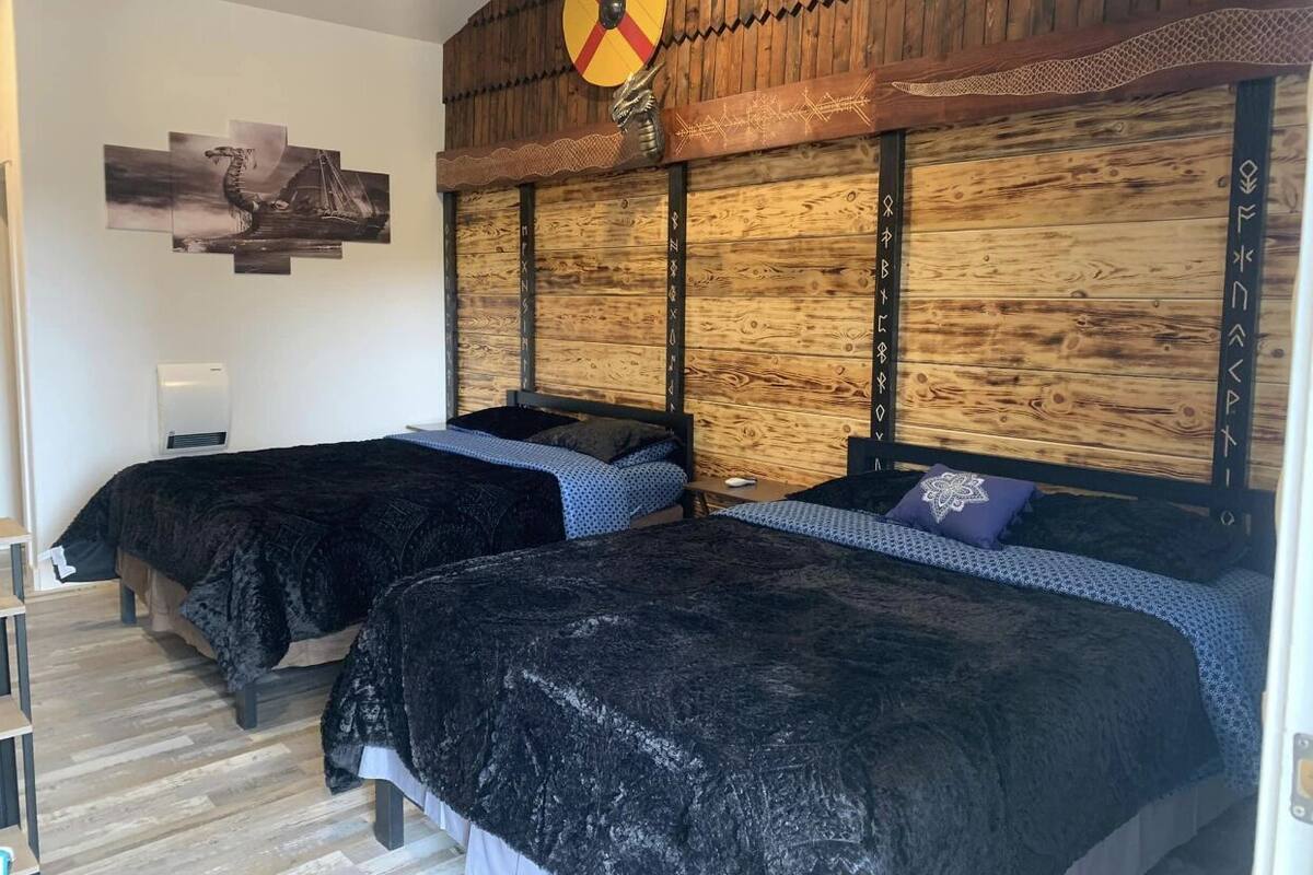 Une chambre avec deux lits, une tête de lit en bois ornée de sculptures, une œuvre d’art accrochée au mur et un ventilateur de plafond.