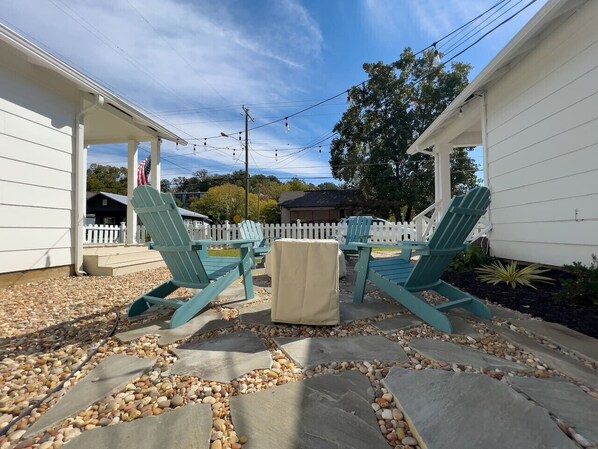 A patio area with Adirondack chairs and a small table, surrounded by a white picket fence and a white building.