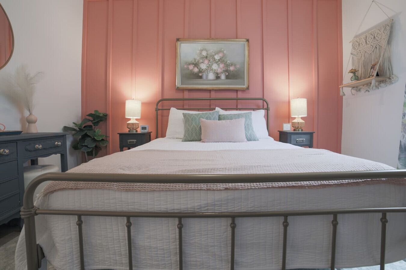 A bedroom with a metal bed frame, two bedside tables with lamps, a dresser, and a wall-mounted macrame piece.