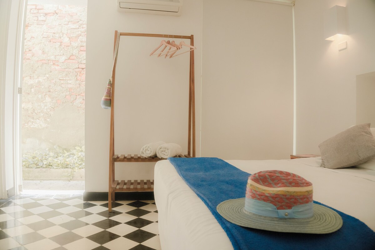 Ein Schlafzimmer mit Schachbrettfußboden, einem Bett mit blauer Decke, einem Hut auf dem Bett, einem hölzernen Wäscheständer mit Handtüchern und einer wandmontierten Lampe.