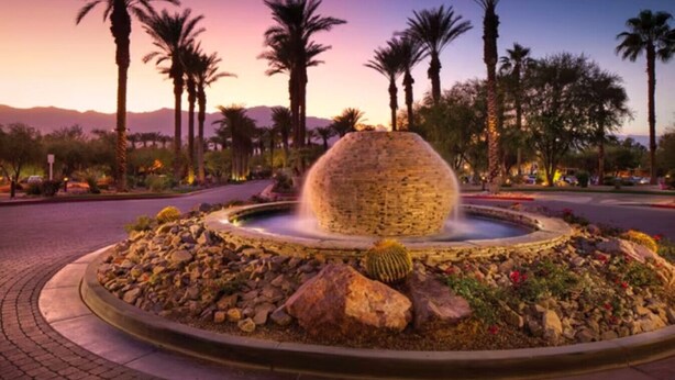 Une fontaine aménagée avec une pièce centrale en pierre sphérique, entourée de rochers et de cactus, le tout se dressant sur un fond de palmiers et d’un ciel au coucher du soleil.