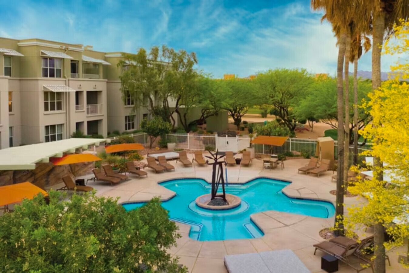 A pool area with lounge chairs and umbrellas, surrounded by greenery and a building with balconies.