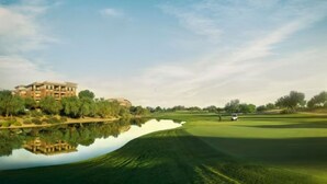 Campo de golf con un cuerpo de agua que refleja el entorno, un edificio al fondo y cielo despejado.