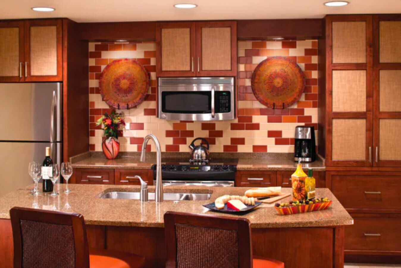 A modern kitchen with a stainless steel refrigerator, a microwave, and a coffee maker. There are two chairs and a dining table with a fruit bowl and a bottle of wine.