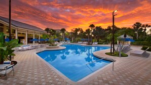 A swimming pool with lounge chairs and umbrellas, surrounded by palm trees and a building with a tiled roof.