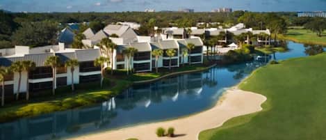 Un campo de golf con un estanque, una zona de arena y edificios modernos con piscina.