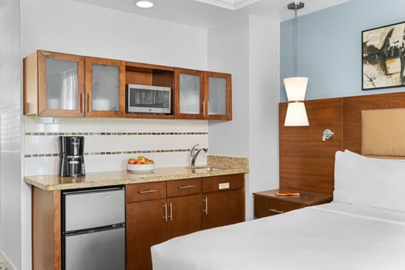 A hotel room with a kitchenette, a bed, a nightstand, a lamp, and a painting on the wall.