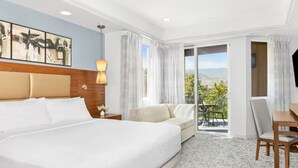 A hotel room with a large bed, a desk, a chair, a lamp, and a balcony with a view of the outdoors.