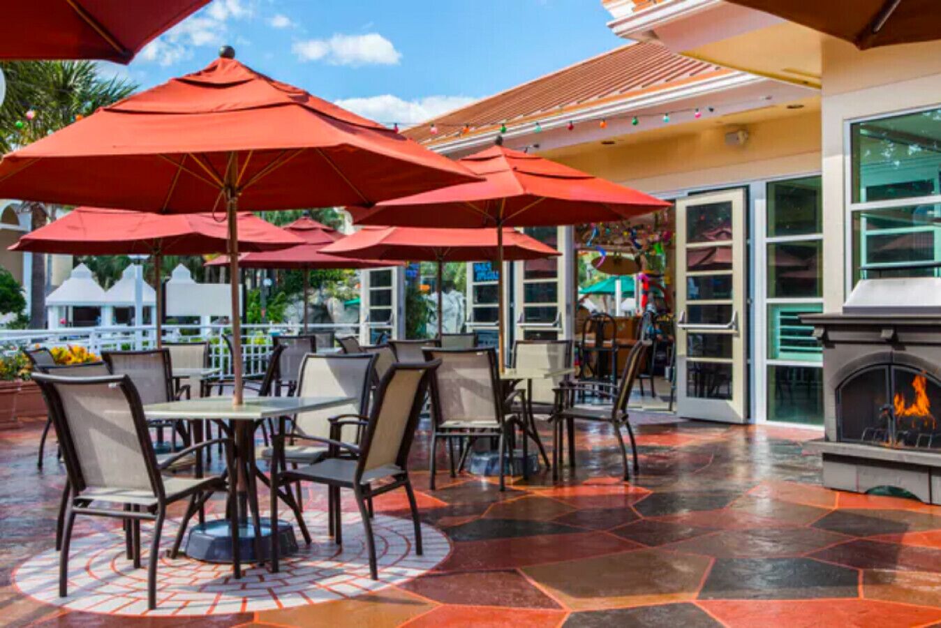 Patio esterno con tavoli e sedie rotonde, ombrelloni rossi e un camino.