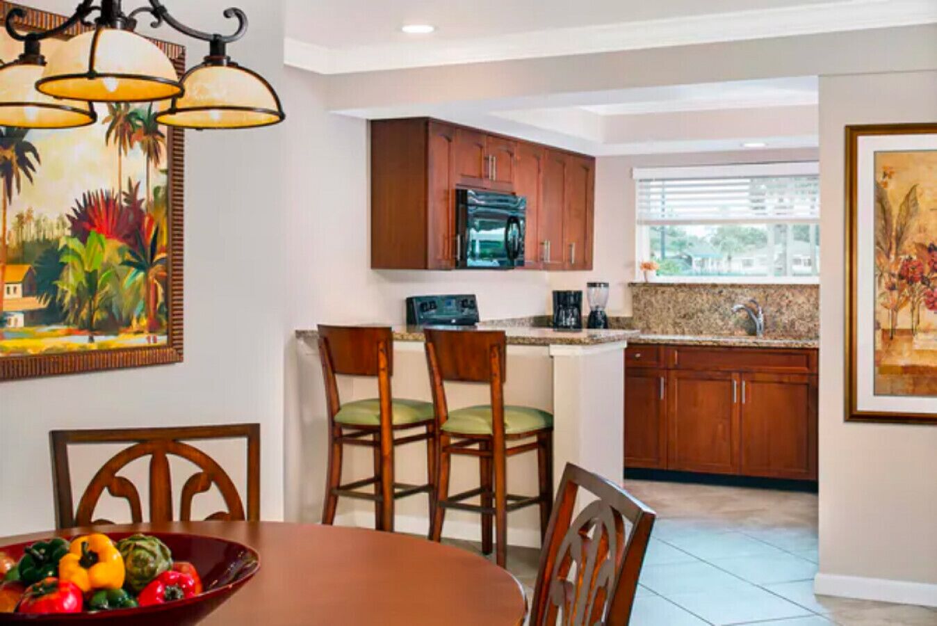 A dining area with a table set for two, a kitchen with wooden cabinets and a microwave, and a window with blinds.
