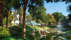 A lush garden with a fountain, palm trees, and a building with a covered walkway. - Sheraton Vistana Resort (1BR) (Orlando)