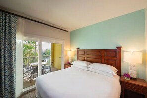 A hotel room with a large bed, two bedside tables with lamps, a balcony with a table and chairs, and a view of greenery outside. - Sheraton Vistana Villages (2BR) (Orlando)