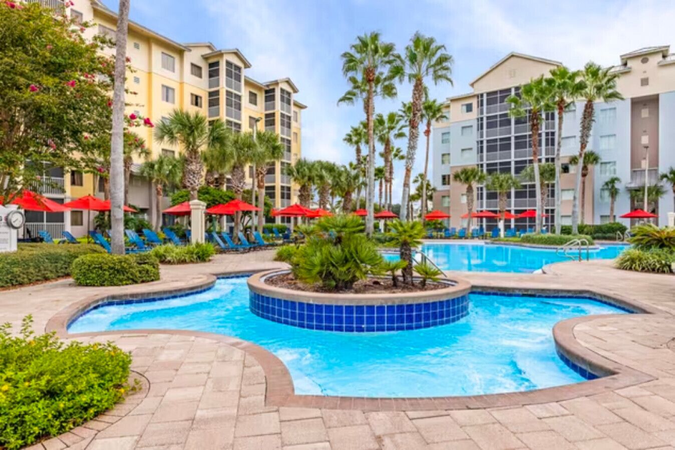 A hotel pool area with palm trees, lounge chairs, and red umbrellas.