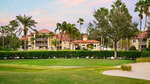 A golf course with a well-maintained lawn, sand bunkers, and a row of multi-story buildings with red roofs and balconies. - Sheraton PGA Vacation Resort (2BR) (Port St. Lucie)