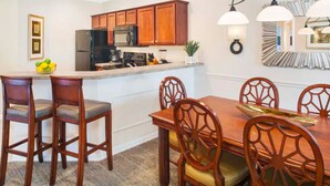 A dining area with a wooden table and chairs, a kitchen with wooden cabinets and appliances, and a decorative wall piece. - Sheraton PGA Vacation Resort (2BR) (Port St. Lucie)