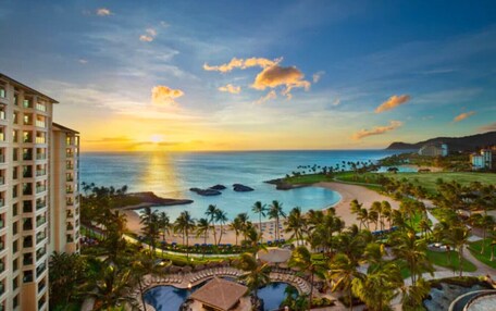 A resort with a pool, palm trees, and a view of the ocean at sunset.