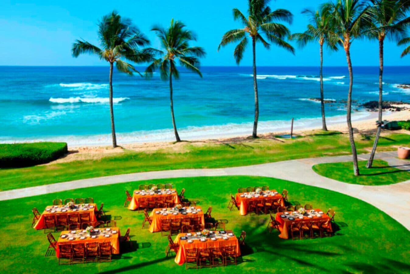 Un área de comedor frente a la playa con mesas preparadas para una comida, palmeras y el océano al fondo.