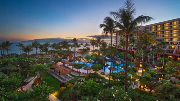 Un resort con un amplio área de piscina, palmeras y vista al océano al atardecer.