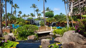 Un resort tropical con una cascada, palmeras, un puente y una piscina rodeada de rocas y lirios de agua.