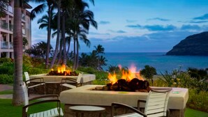 Un espacio al aire libre con zona de estar y chimenea, rodeado de palmeras y vistas a la costa.