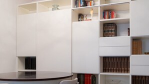 Una habitación con una mesa redonda y sillas, una estantería blanca con libros, y un objeto decorativo en la repisa superior.