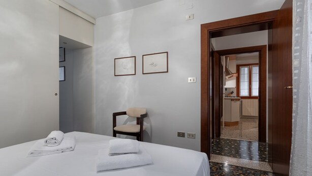 Habitación de hotel con cama, una silla, dos cuadros en la pared y una puerta que da a una zona de cocina.