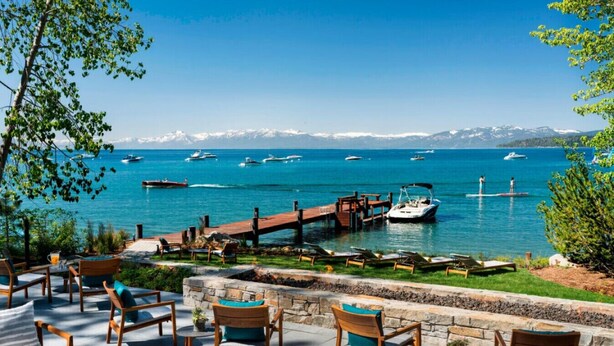 Une terrasse au bord de l’eau, avec des chaises et des tables en bois, un quai avec des bateaux, et une vue dégagée sur la mer et les montagnes en arrière-plan.