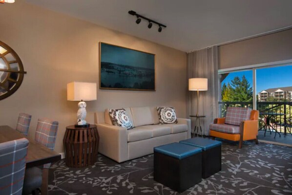 A hotel room with a sofa, two ottomans, a table, and a chair. There is a painting on the wall and a window with a view of buildings.