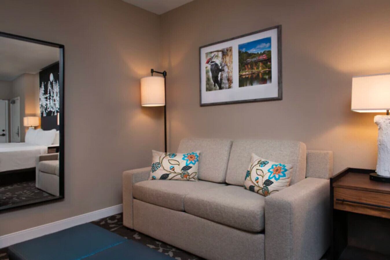 A hotel room with a beige sofa, two floral pillows, a framed picture above the sofa, a standing lamp, and a bed with a nightstand.