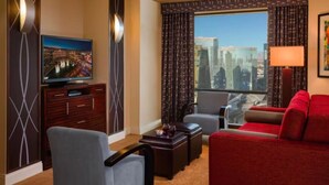 A hotel room with a city view, a flat-screen TV, a red sofa, a grey armchair, and a small table with a lamp. - Marriott's Grand Chateau (3BR) (Las Vegas)