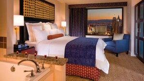 A hotel room with a large bed, a view of a cityscape, a sitting area with a blue sofa, and a bathroom with a sink and mirror. - Marriott's Grand Chateau (3BR) (Las Vegas)