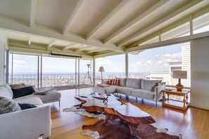 Un salon spacieux avec un plafond haut, de grandes fenêtres offrant une vue sur la ville, une table basse en cuir et un canapé.
