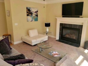 A living room with a beige sofa, a glass coffee table with magazines, a fireplace, and a painting on the wall.