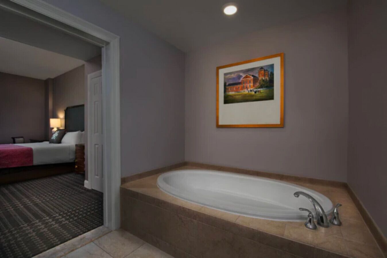 Quarto de hotel com uma banheira grande, uma cama com manta vermelha e um quadro na parede.