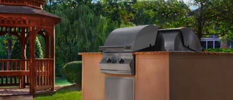 An outdoor barbecue grill with a chimney, a wooden gazebo, and a well-maintained garden in the background.