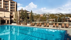 En swimmingpool med liggestole, omgivet af bygninger og sneklædte bjerge.
