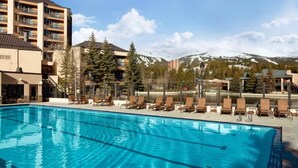 Piscina con sillas de descanso, rodeada de edificios y montañas nevadas.