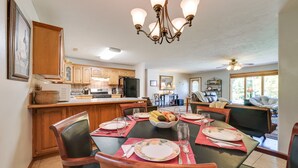 A dining area with a large table set for six, surrounded by wooden chairs. A chandelier hangs above, and a kitchen is visible in the background. - Game Room & Deck: Serene Ava Countryside Home (Ava)