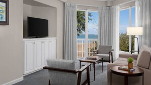 A modern hotel room with a flat-screen TV, a sofa, a chair, a small table with books, and a view of the sea through large windows.