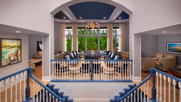 A spacious living area with a high ceiling, large windows, and a staircase leading to an upper floor.