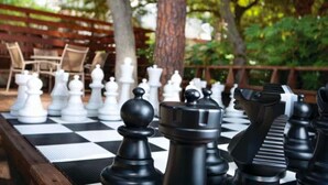 A chessboard with pieces set up for a game, surrounded by outdoor furniture and trees.