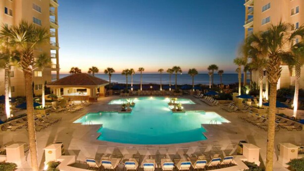 A resort pool with multiple sections, surrounded by palm trees and lounge chairs, with a view of the ocean and a building in the background. - OceanWatch (2 Bedroom) (Myrtle Beach)