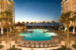 A resort pool with multiple sections, surrounded by palm trees and lounge chairs, with a view of the ocean and a building in the background.