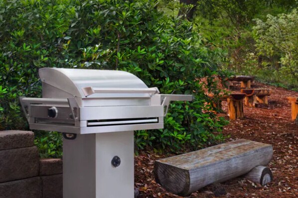 A barbecue grill in a garden setting with a wooden table and benches in the background.